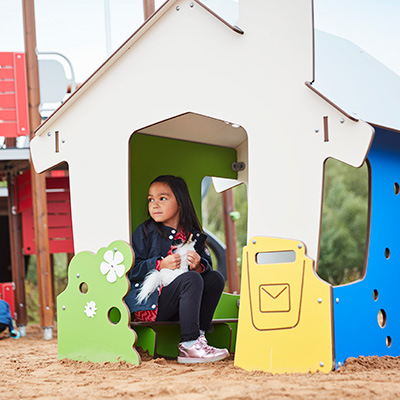 A young girl is sitting inside a colorful playhouse in a playground, she is looking to the side.